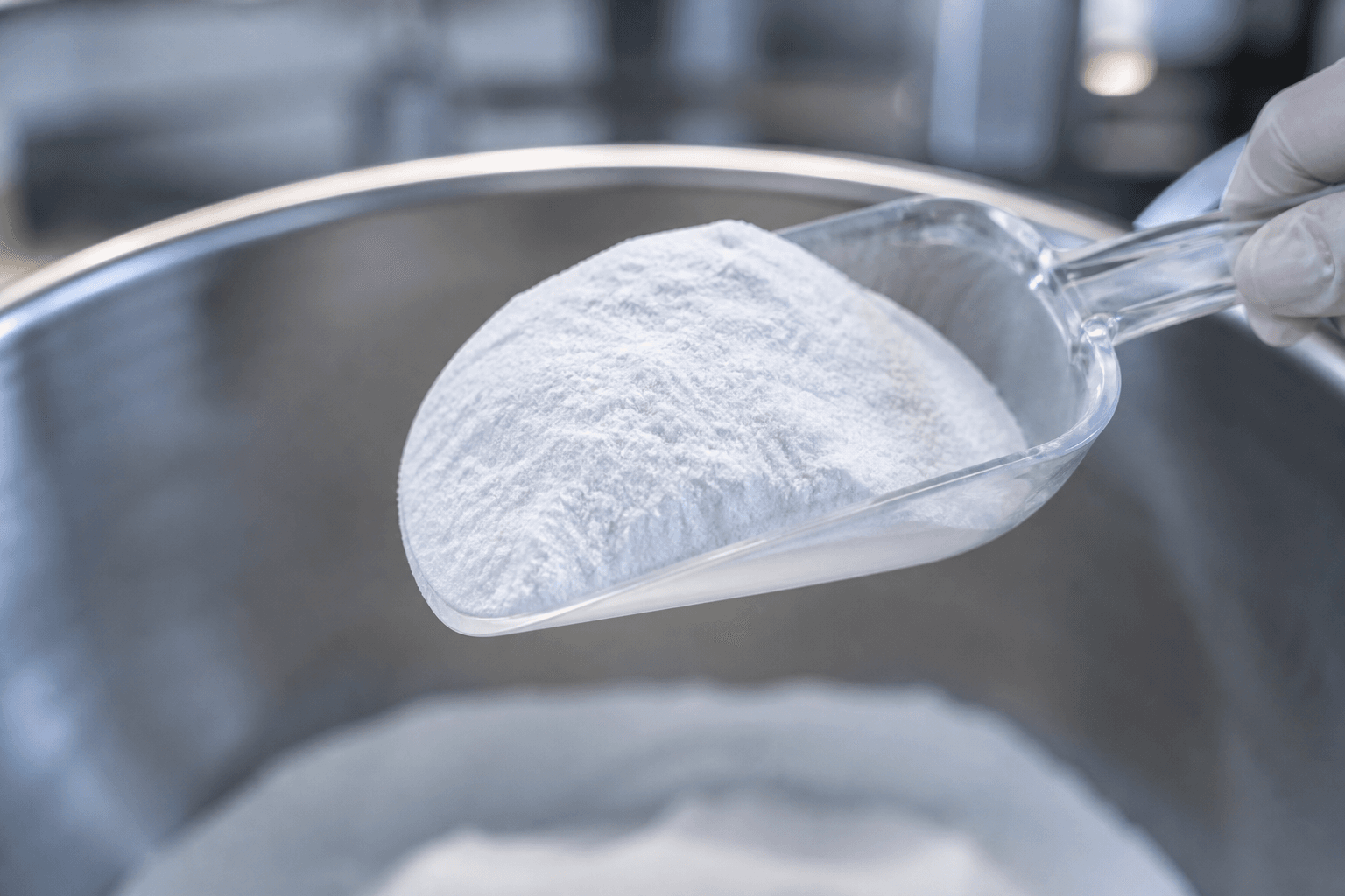 Gloved hand holding a clear scoop of fine white powder over a stainless steel container