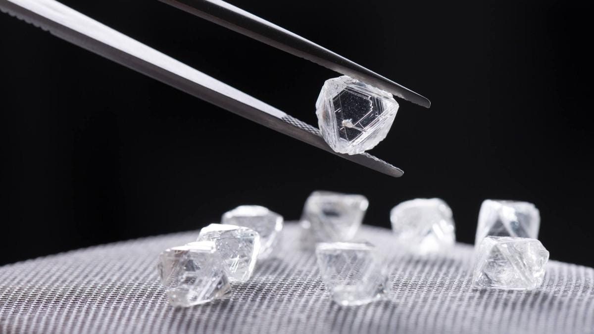 Clear rough diamond held in tweezers above more stones on a grey mesh surface