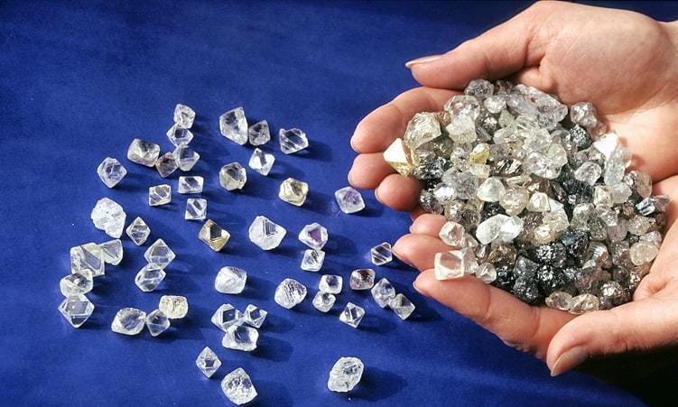 Hands holding mixed clear, yellow, and dark rough diamonds on blue fabric